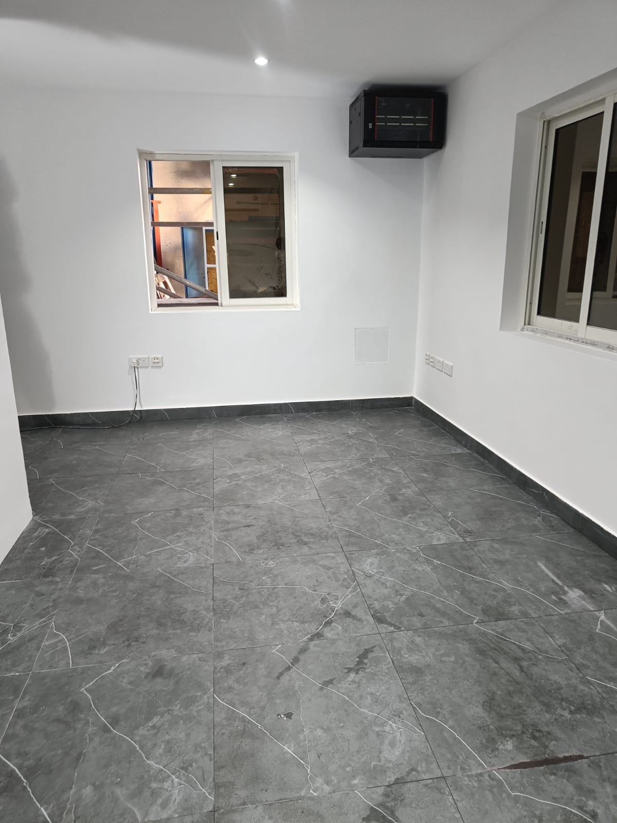 Empty renovated office room with white walls, marble-pattern floor tiles, windows, and wall-mounted data cabinet